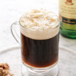 Glass mug of homemade Irish coffee topped with whipped cream on a marble countertop with Irish whiskey in the background