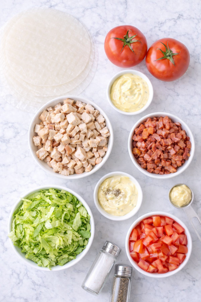 flat lay of ingredients for club sandwich rice paper spring rolls with turkey bacon lettuce tomato and rice paper wrappers on marble background