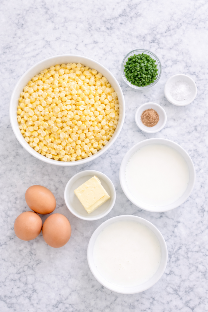 Flat lay of corn pudding ingredients including fresh corn kernels, eggs, milk, cream, butter, chives, salt, nutmeg, and white pepper on a marble countertop