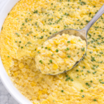 Spoon scooping creamy homemade corn pudding from a white baking dish, bird’s-eye view on marble countertop