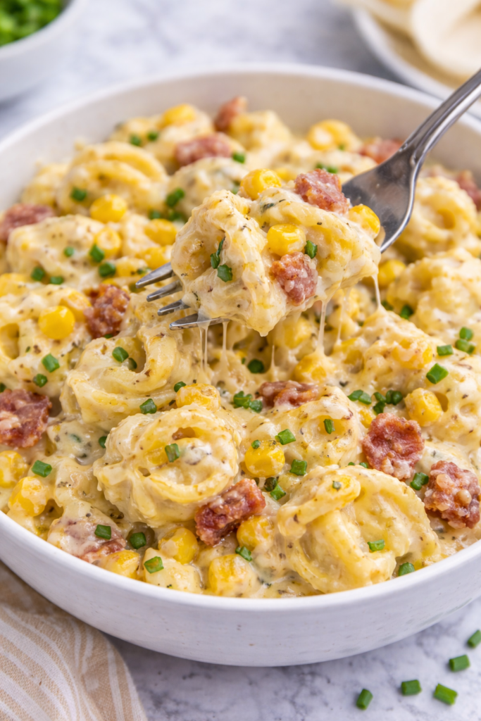fork scooping creamy corn and bacon tortellini from white bowl on marble counter