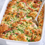 Creamy Dijon croissant breakfast casserole with ham and Swiss cheese baked in a white casserole dish on a marble counter, topped with chopped chives and parsley with a serving spoon tucked into the casserole.