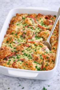 Creamy Dijon croissant breakfast casserole with ham and Swiss cheese baked in a white casserole dish on a marble counter, topped with chopped chives and parsley with a serving spoon tucked into the casserole.
