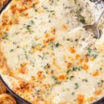 creamy garlic herb artichoke dip baked in skillet with melted cheese and toasted baguette slices