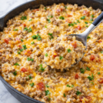 Creamy ground beef and rice skillet with melted cheese, diced tomatoes, and a spoon scooping a serving from a cast iron pan on a marble countertop