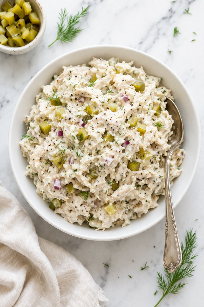 creamy pickle chicken salad with dill pickles and red onion in white bowl on marble counter with spoon