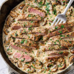Creamy steak marsala and mushrooms in a black skillet with sliced steak, a rich marsala cream sauce, sautéed mushrooms, fresh parsley, and a serving fork on a white marble countertop.