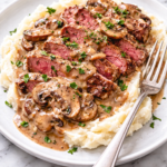 Sliced steak marsala with creamy mushroom gravy served over mashed potatoes on a white plate with a fork on a white marble countertop.
