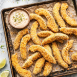 Crispy baked avocado fries on a rustic baking sheet with wrinkled parchment paper, served with lime ranch dipping sauce on a marble countertop.
