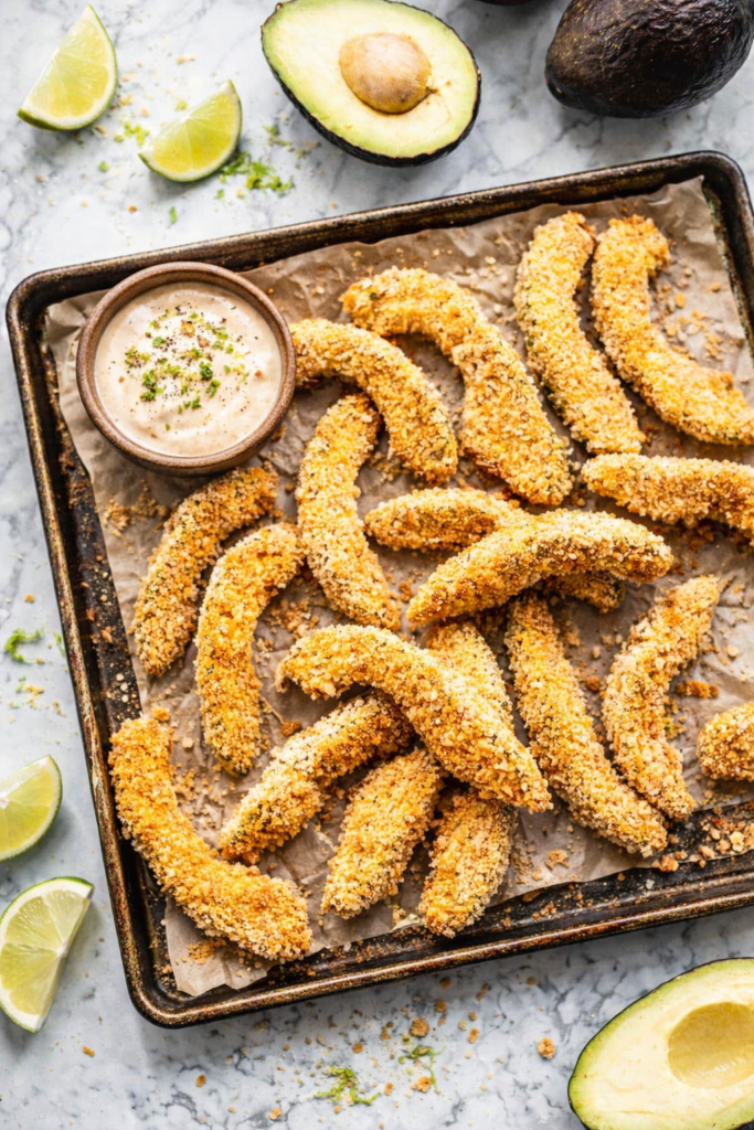 Crispy baked avocado fries on a rustic baking sheet with wrinkled parchment paper, served with lime ranch dipping sauce on a marble countertop.