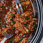Crockpot shredded beef in grape jelly and ketchup sauce being pulled apart with forks in a slow cooker with parsley garnish