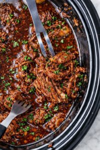 Crockpot shredded beef in grape jelly and ketchup sauce being pulled apart with forks in a slow cooker with parsley garnish