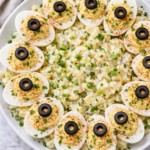 Deviled egg potato salad topped with paprika, parsley, and black olive slices served on a white platter on a marble countertop