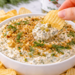 Crinkle cut potato chip being dipped into creamy dill pickle dip topped with toasted breadcrumbs and fresh dill in a white bowl on a marble countertop.