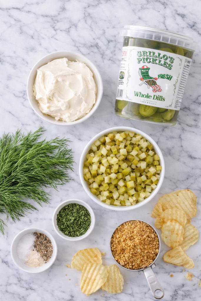 Flat lay of ingredients for dill pickle dip including cream cheese, chopped pickles, fresh dill, seasonings, breadcrumbs, and crinkle cut potato chips on a marble countertop.