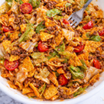 Fork lifting a bite of Dorito taco salad with lettuce, ground beef, cherry tomatoes, shredded cheese, and Catalina dressing in a white bowl on a marble counter