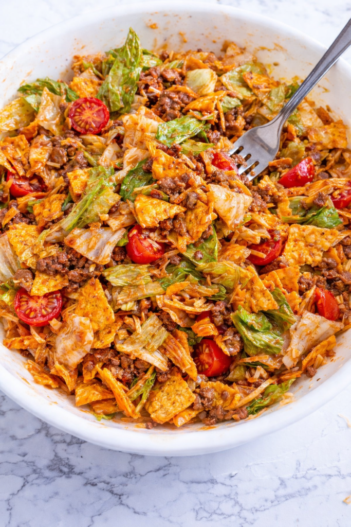 Fork lifting a bite of Dorito taco salad with lettuce, ground beef, cherry tomatoes, shredded cheese, and Catalina dressing in a white bowl on a marble counter