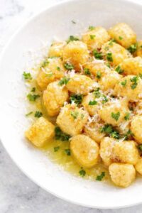 Easy homemade potato gnocchi tossed in butter with parmesan cheese and fresh parsley in a white bowl on a marble counter