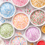 Flat lay of homemade fancy sugar mixes in white bowls with colorful decorating sugar blended into granulated sugar on a marble countertop