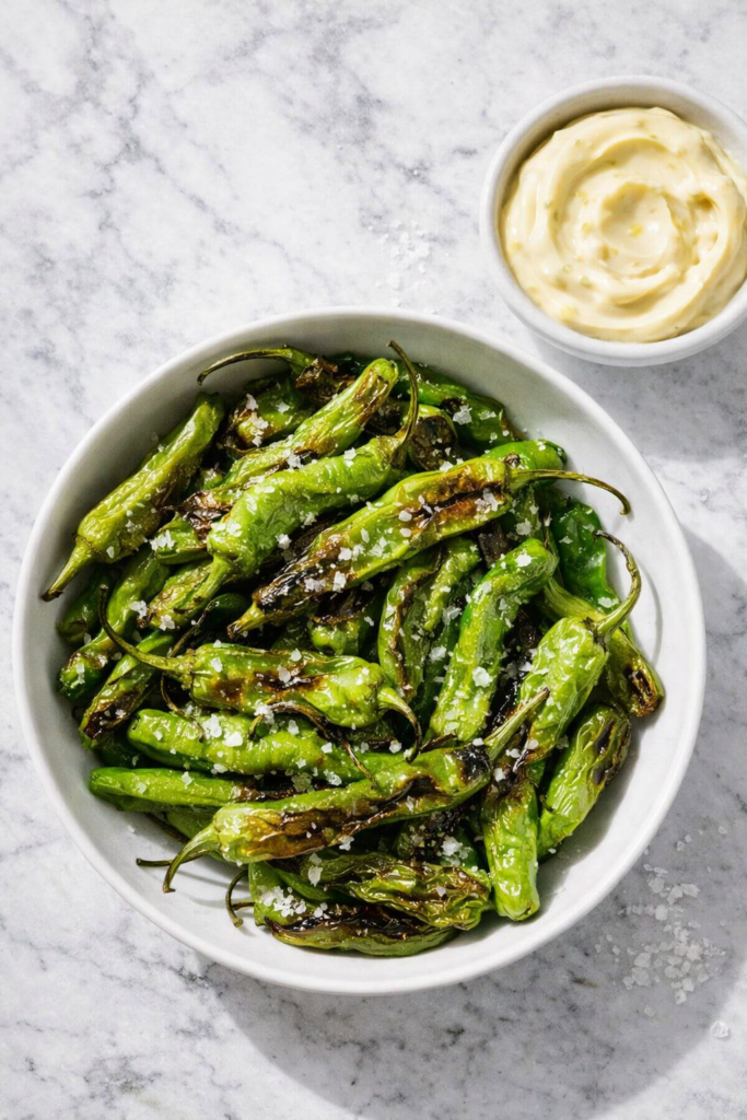 Blistered shishito peppers in a white bowl on a marble counter with flaky sea salt and a side of creamy garlic aioli