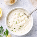 Creamy garlic aioli in a white bowl on a marble countertop with fresh garlic cloves, lemon, and parsley