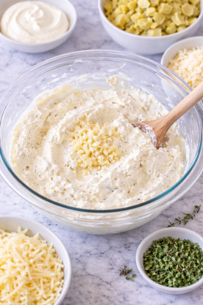 creamy garlic herb artichoke dip base being mixed in glass bowl with cream cheese sour cream and garlic