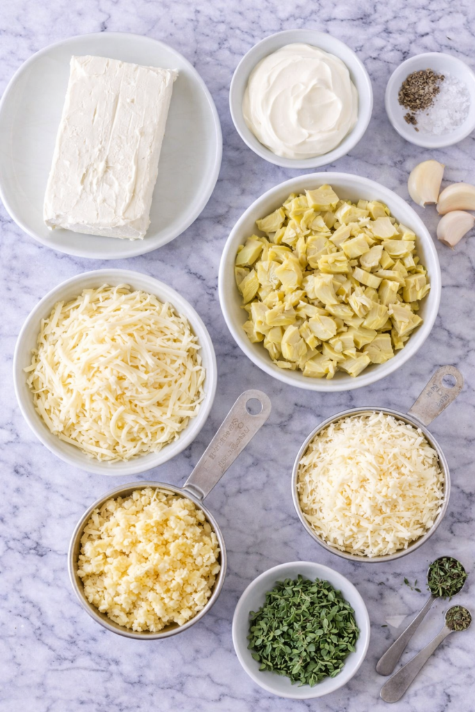 flat lay of ingredients for garlic herb artichoke dip including cream cheese sour cream artichokes mozzarella parmesan garlic and fresh herbs