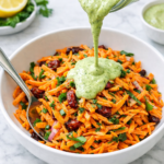Alt Text: Green goddess carrot slaw with dried cranberries and creamy herb dressing being poured over shredded carrots in a white bowl on a marble countertop