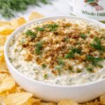 Creamy dill pickle dip topped with toasted breadcrumbs and fresh dill served in a white bowl with crinkle cut potato chips on a marble countertop with a container of Grillo’s Pickles in the background.