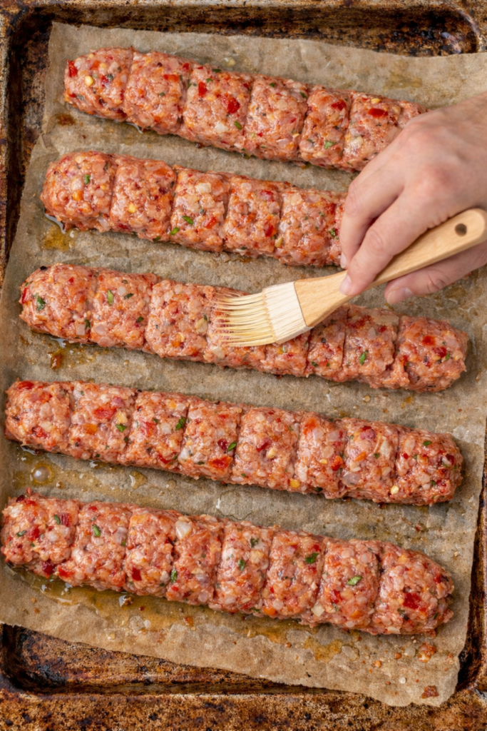Raw ground chicken shawarma oven kabobs on parchment lined baking sheet being brushed with oil using a pastry brush