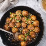 Gyoza meatballs pan-seared in a cast iron skillet with soy ginger glaze, garnished with green onions and sesame seeds on a marble countertop