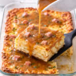Thin red eye gravy being poured over a slice of ham and egg breakfast casserole in a glass baking dish on a marble countertop