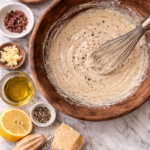 Homemade Caesar dressing being whisked in a wooden bowl on a marble countertop with garlic, anchovies, lemon, olive oil, and Parmesan.