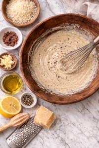 Homemade Caesar dressing being whisked in a wooden bowl on a marble countertop with garlic, anchovies, lemon, olive oil, and Parmesan.