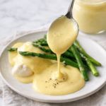 Homemade hollandaise sauce drizzled over asparagus on a neutral ceramic plate with a mason jar of sauce on a marble countertop