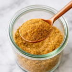 Copper spoon scooping homemade shawarma seasoning from a glass jar on a white marble countertop