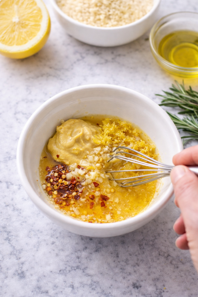 Whisking honey mustard glaze with Dijon mustard, lemon zest, garlic, and honey in a white bowl on a marble countertop