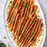 Honey roasted carrots arranged on a white oval platter over creamy whipped feta, finished with chopped parsley and a drizzle of honey on a marble countertop.