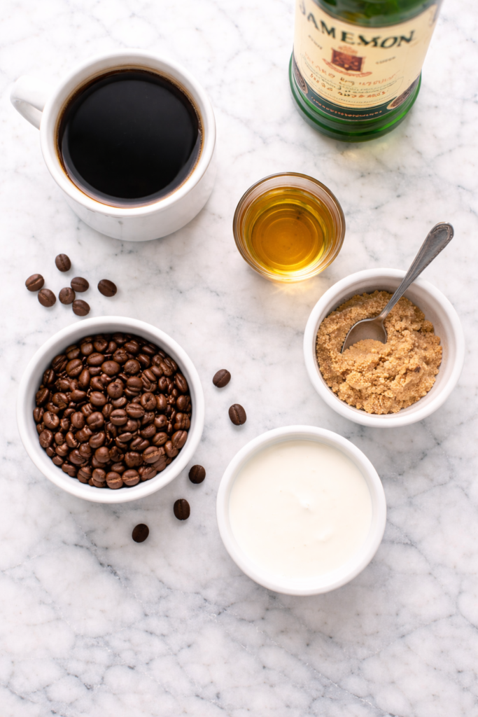 Flat lay of Irish coffee ingredients including brewed coffee, Irish whiskey, brown sugar, cream, and coffee beans on a marble countertop