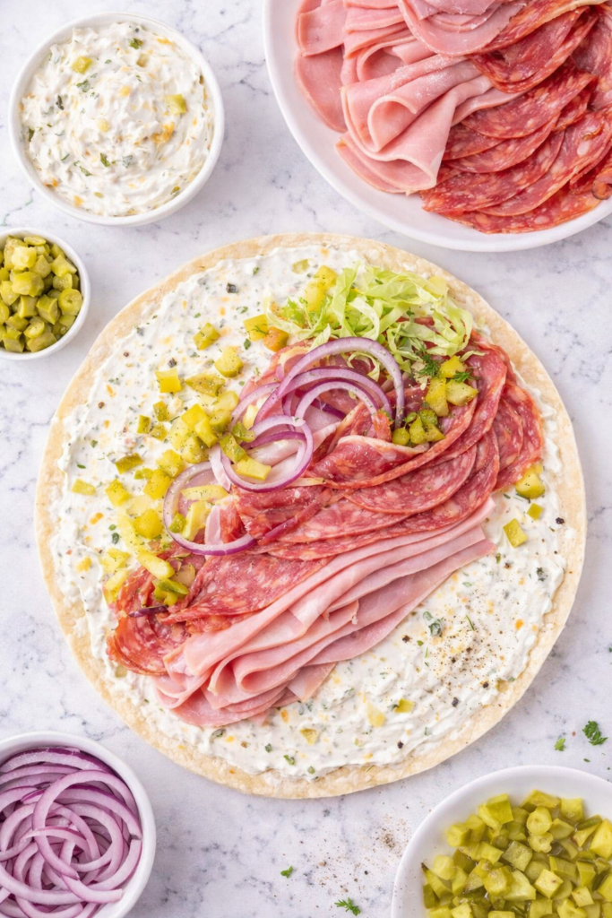 tortilla with pickle dip ham salami and capicola being layered before rolling into italian grinder pinwheel sandwiches