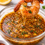 Hand holding a jumbo shrimp by the tail dipping it into a bowl of garlic butter seafood boil sauce with garlic and Cajun spices.