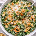 Fork lifting kale and carrot Caesar salad with creamy dressing, shaved carrots, Parmesan cheese, and crushed croutons in a white bowl on a marble counter
