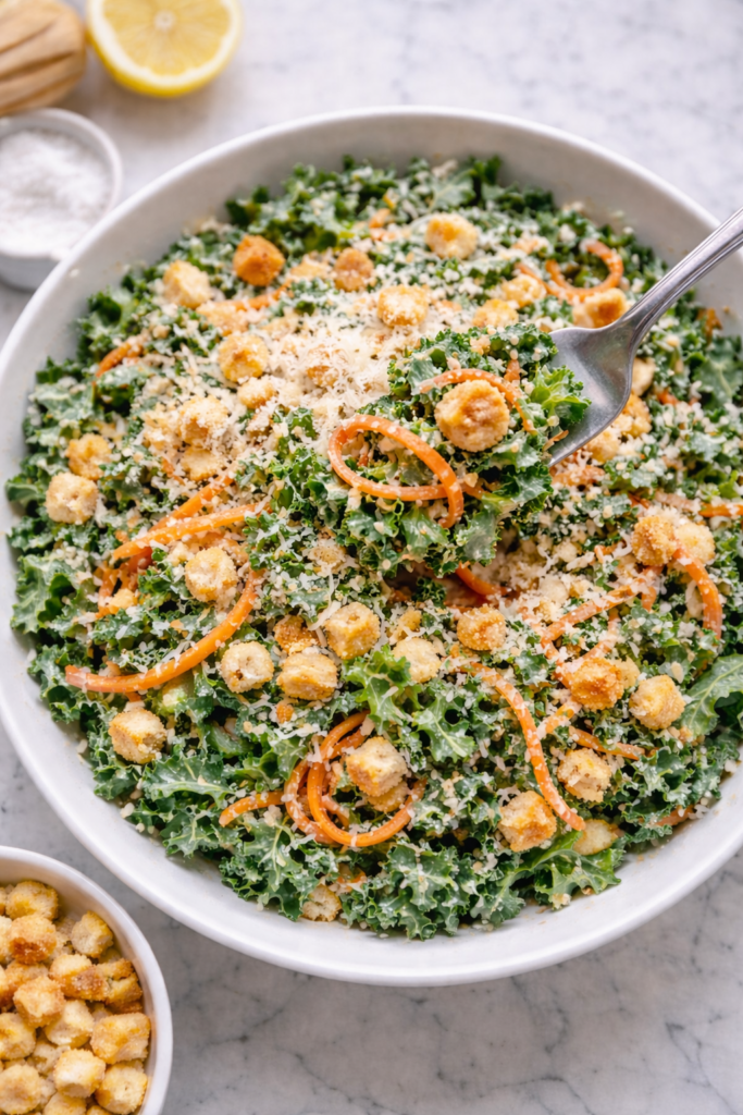 Fork lifting kale and carrot Caesar salad with creamy dressing, shaved carrots, Parmesan cheese, and crushed croutons in a white bowl on a marble counter