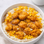 Homemade KFC Famous Bowl copycat with mashed potatoes, crispy fried chicken, sweet corn, brown gravy and melted cheddar cheese in a white bowl on a marble counter