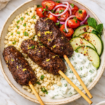 Lebanese kafta kabobs served in a rustic ceramic bowl with pearl couscous, creamy yogurt sauce, marinated tomatoes, red onion, and fresh cucumbers on a marble countertop.
