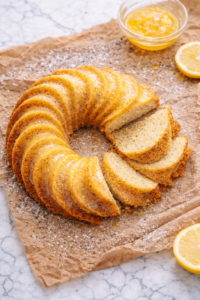 Lemon poppy seed cornmeal bundt cake with lemon glaze on brown wrinkled parchment paper over marble countertop, dusted with powdered sugar and scattered poppy seeds