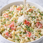 Lobster roll pasta salad in a white bowl with creamy dressing being poured over large chunks of lobster, garnished with chives and parsley on a marble counter