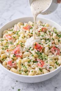 Lobster roll pasta salad in a white bowl with creamy dressing being poured over large chunks of lobster, garnished with chives and parsley on a marble counter