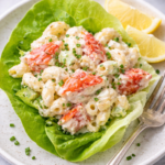 Lobster roll pasta salad served in a butter lettuce cup with large lobster chunks, creamy dressing, and herbs on a white plate with a fork