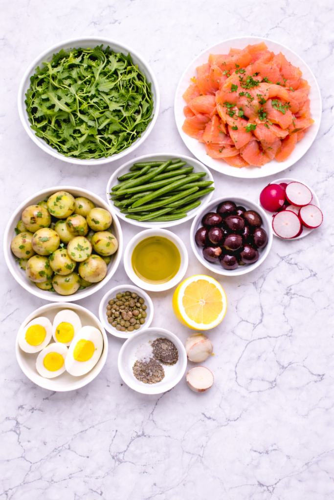 Flat lay of ingredients for a lox Niçoise salad including smoked salmon, potatoes, green beans, olives, eggs, tomatoes, capers, lemon, and arugula on a marble counter